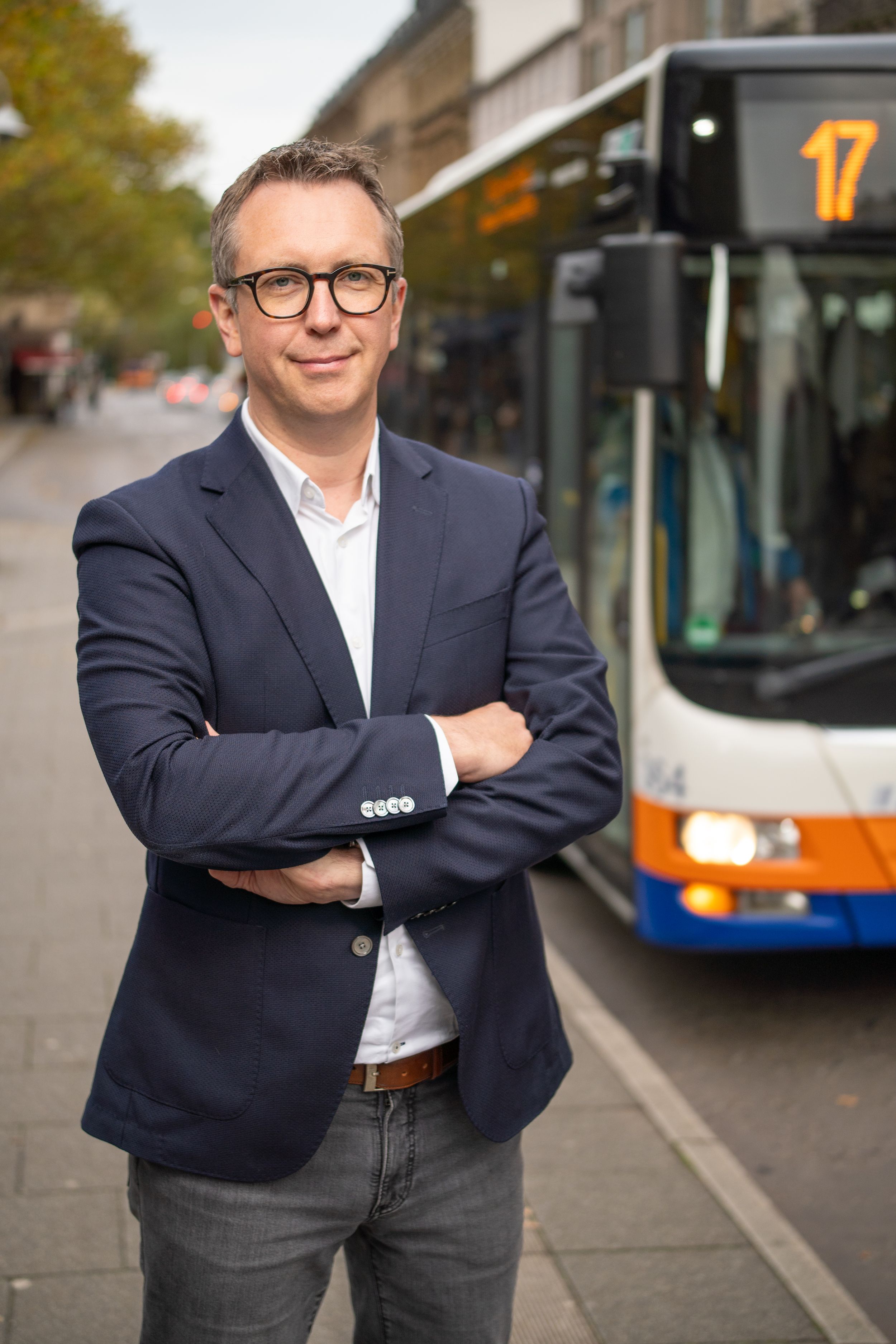 Professional in a navy blazer and glasses poses confidently near city buses on an urban street.