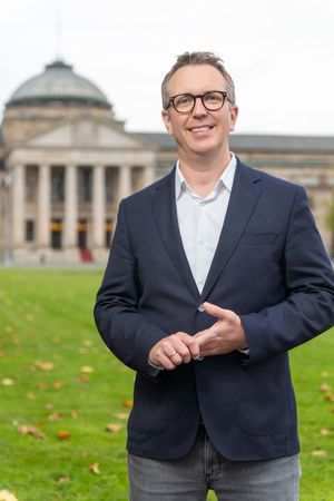 Professional portrait of someone in a navy blazer and glasses standing on a grassy lawn with buildings in background.