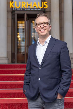 Professional portrait of someone in a navy blazer and white shirt standing in front of a Kurhaus building entrance.