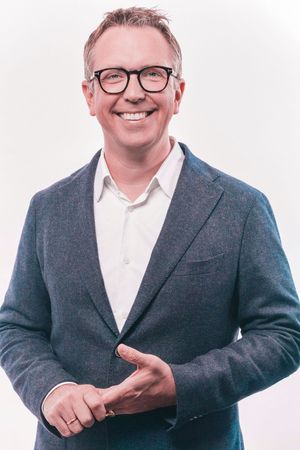 Professional headshot of a person wearing glasses, a white dress shirt and gray blazer, smiling warmly against light background.