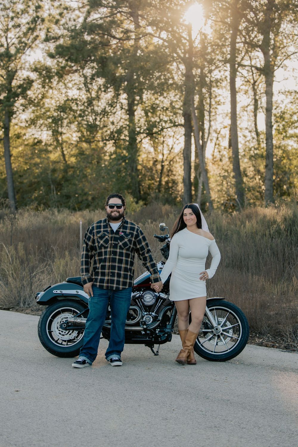 Couple posing with a black motorcycle on a rural road at golden hour surrounded by tall trees.