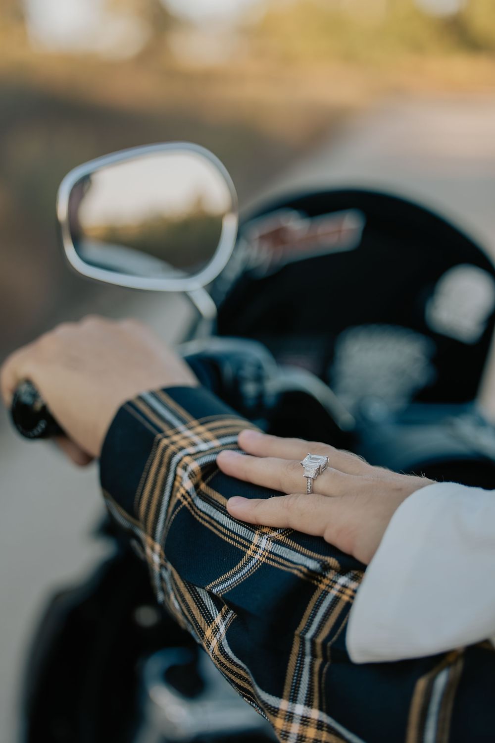 Close-up of a woman's hand wearing a diamond engagement ring resting on a plaid jacket sleeve near a motorcycle mirror.