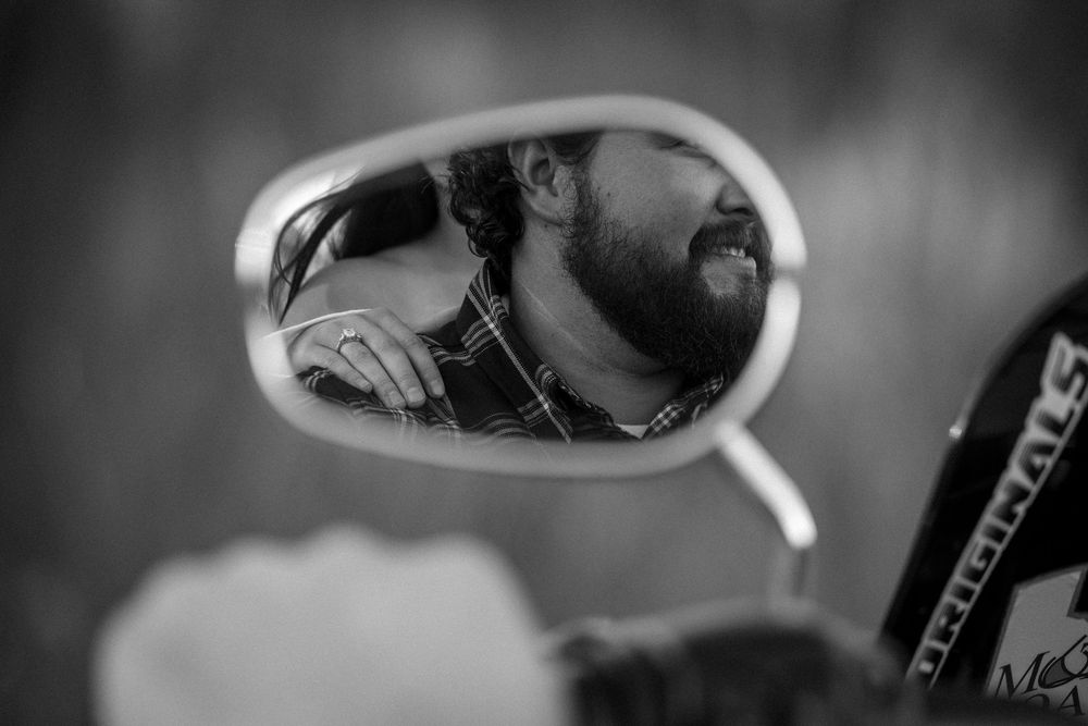 Black and white photo of a bearded man with sunglasses reflected in a motorcycle side mirror.