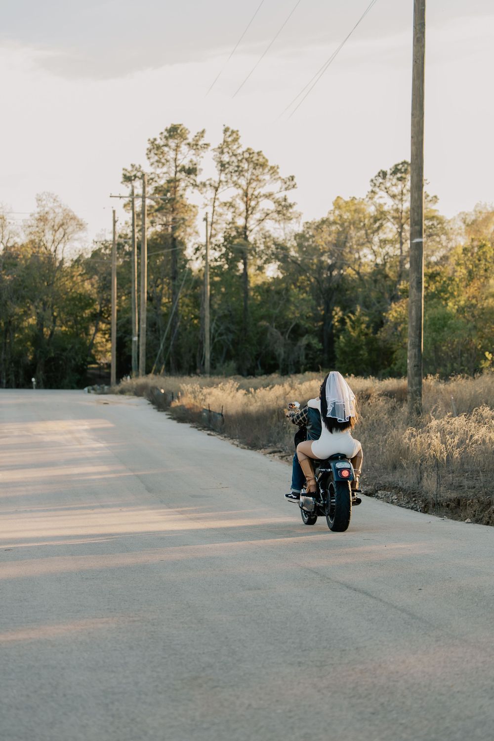 Couple riding a motorcycle down a quiet rural road at sunset with trees lining the path.