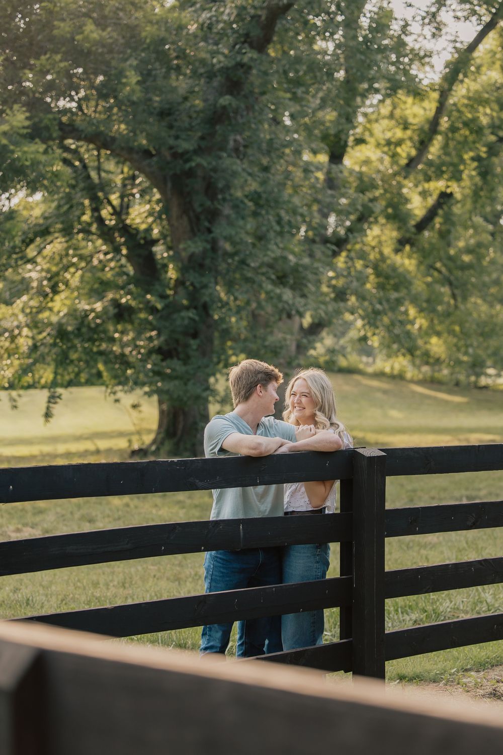 Young couple gazes lovingly at each other leaning on a black wooden farm fence.