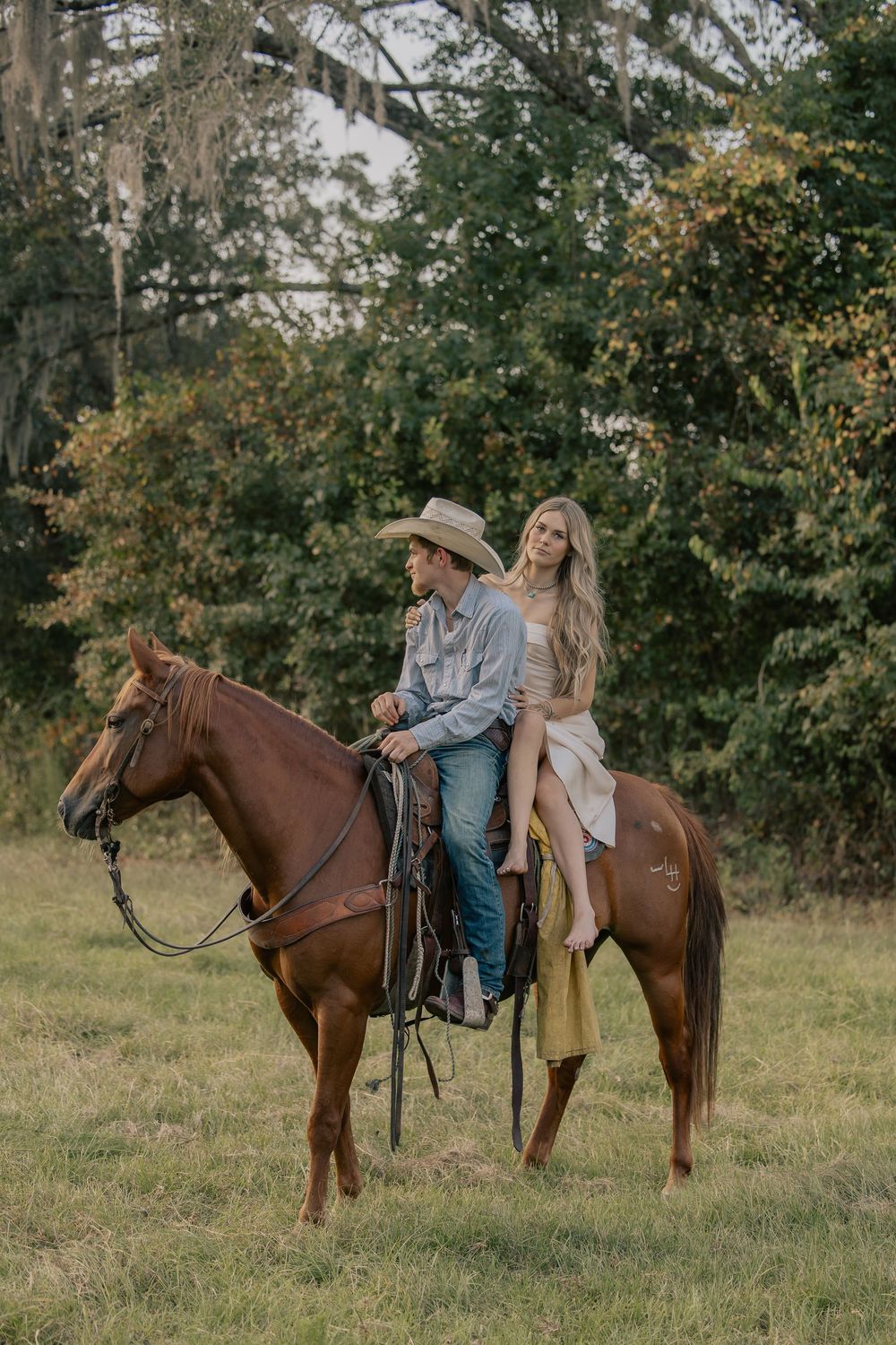 Couple riding a chestnut horse together in a lush green pasture near trees.