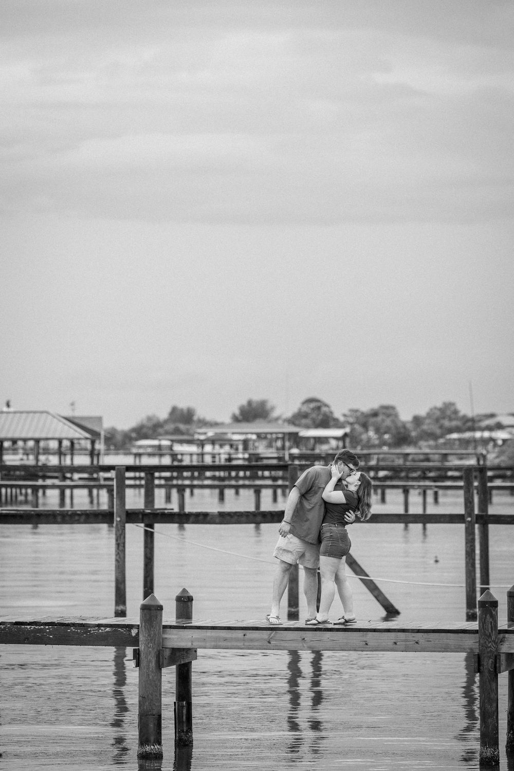 Black and white photo of a couple embracing on a wooden pier with marina in background.