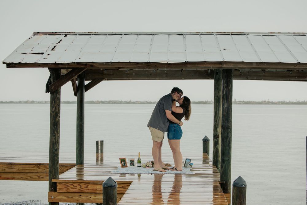 Newly engaged couple embraces and kisses on a rainy wooden dock overlooking a calm bay.