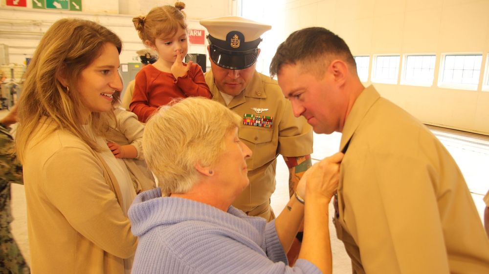 Family-centric Chief pinning ceremony photography showing a mother attaching gold anchors to a Sailor's collar inside an aviation hangar.