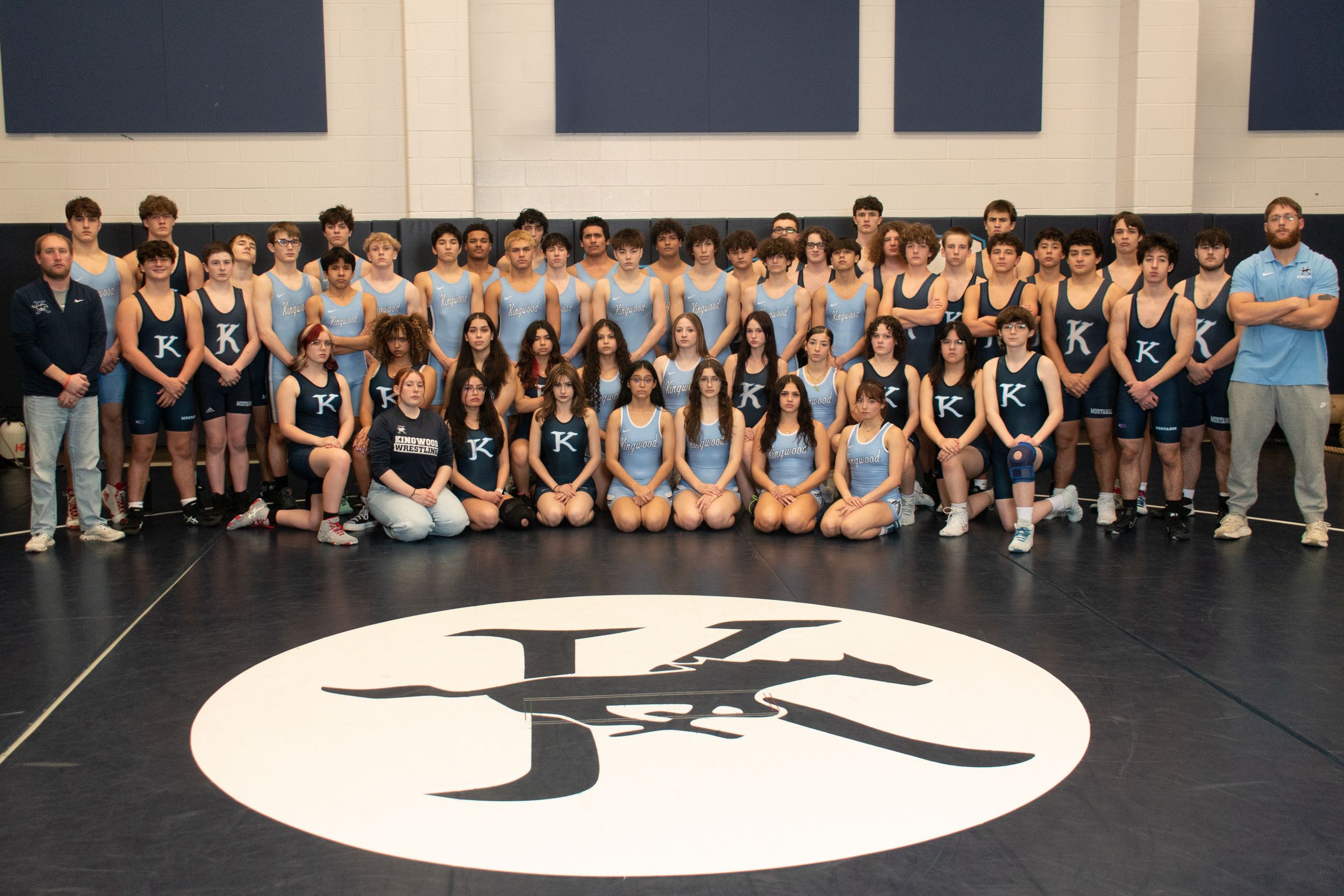 Large group of students and coaches pose together on indoor wrestling mat with team logo.