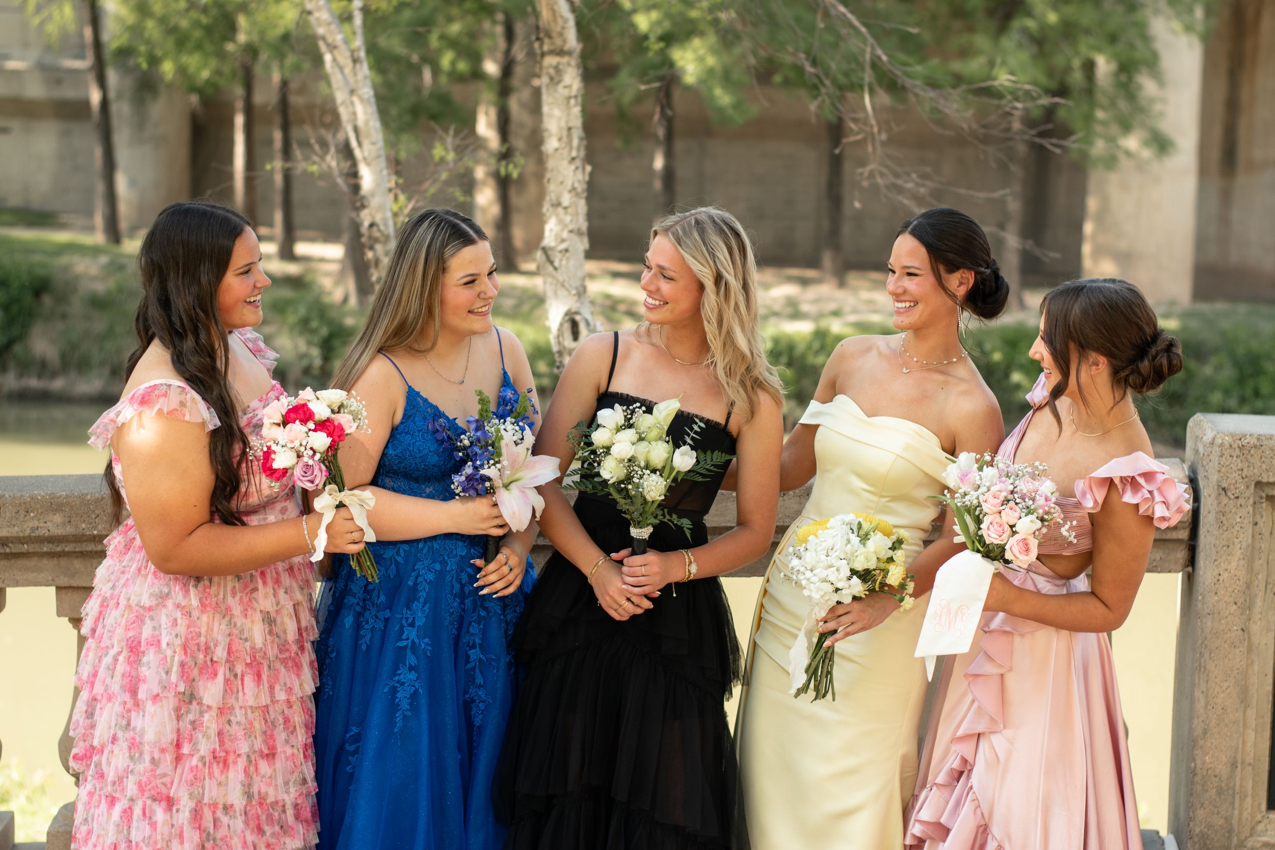 A group of women in elegant formal dresses stand together outdoors holding bouquets during a wedding celebration.