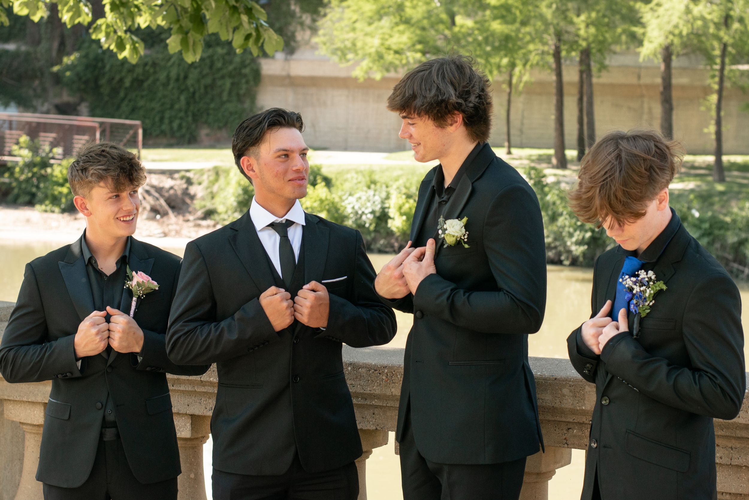 Four people in formal black suits and boutonnières stand together outdoors showing playful gestures.