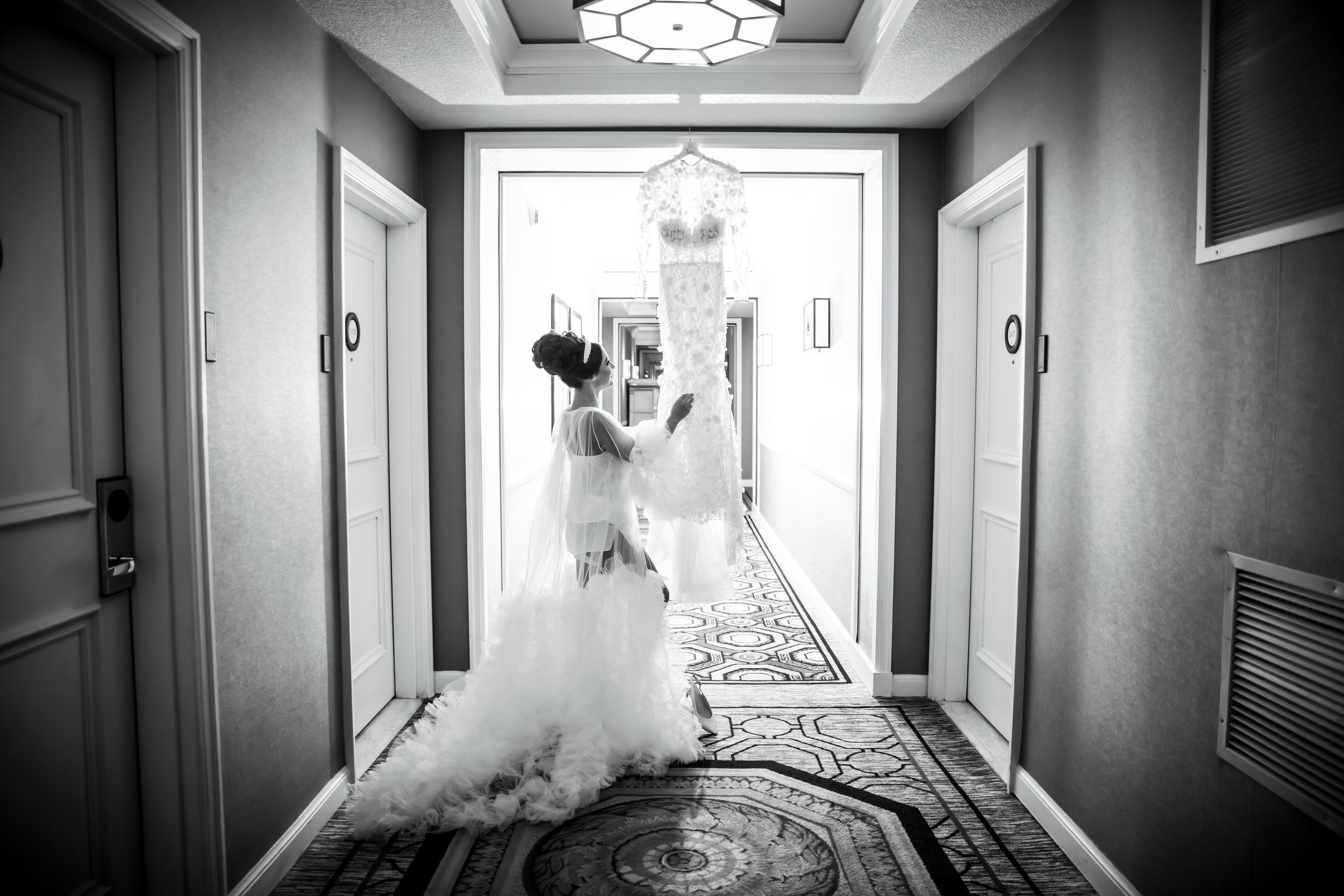 A bride in a flowing white gown walks down an elegant hotel hallway in black and white photography.