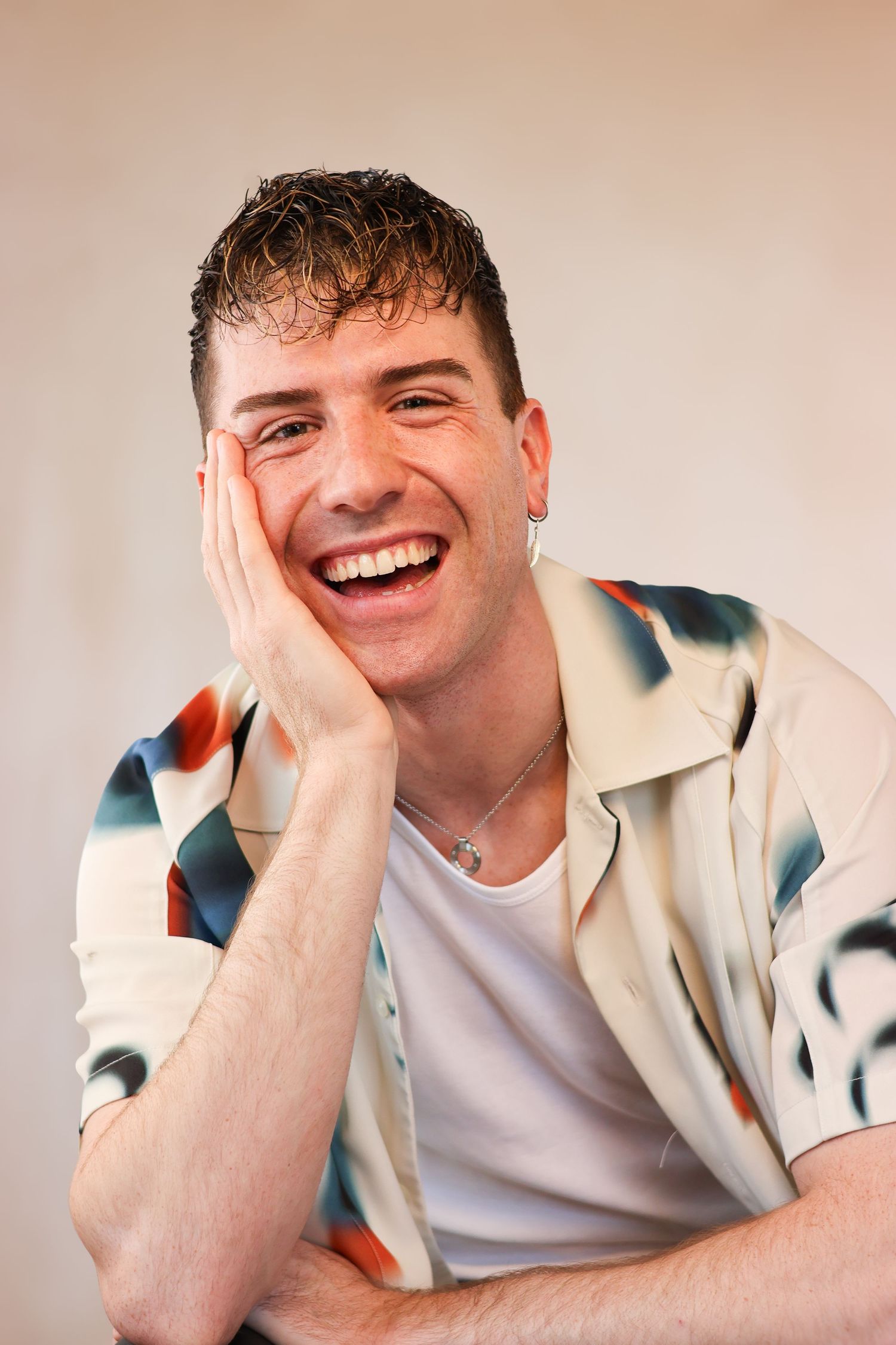 Young person wearing a patterned button-up shirt laughing joyfully with hand on cheek against neutral background.