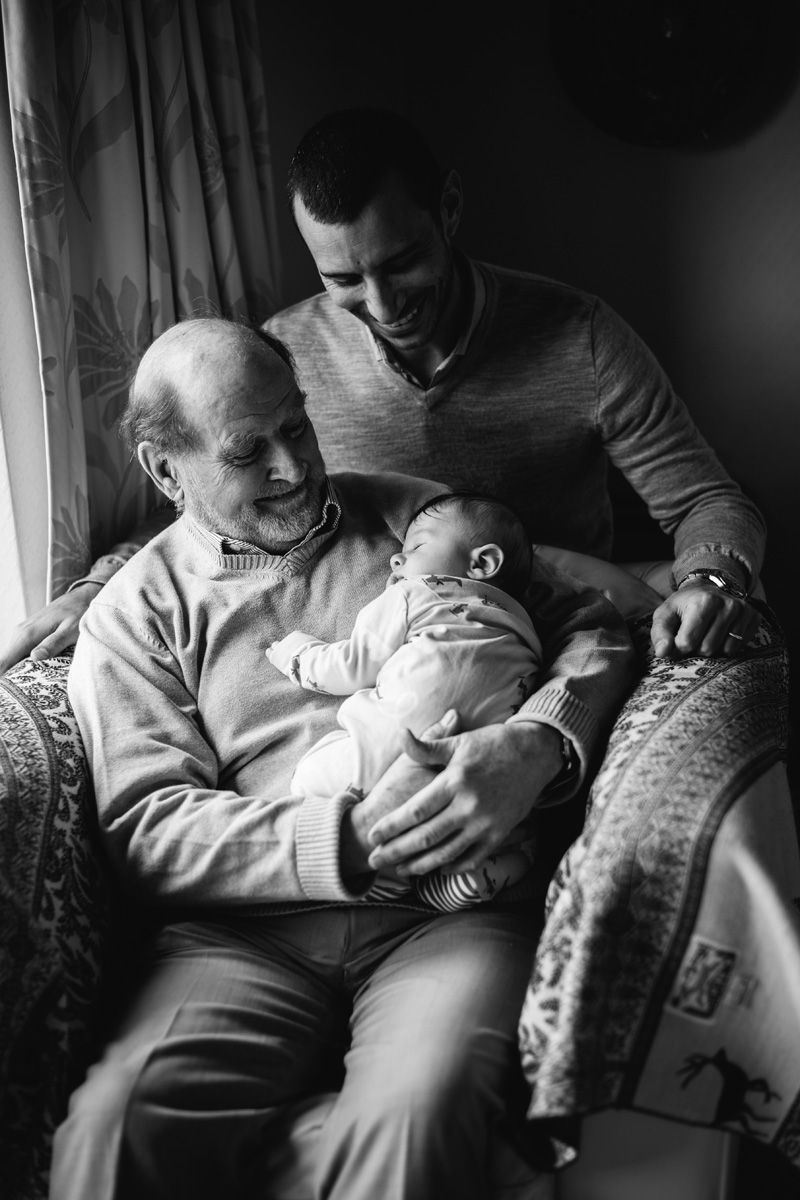 Three-generation family portrait with a grandfather holding a sleeping newborn and the father smiling beside them.