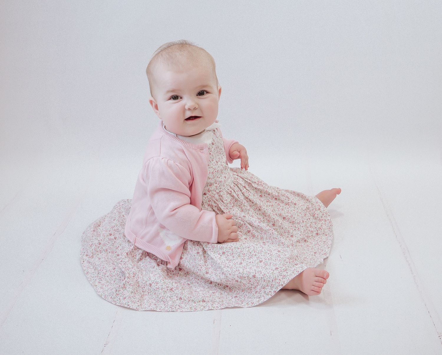 Small baby wearing pink sweater sitting on white floor with floral print dress. Early years pop up photo shoot