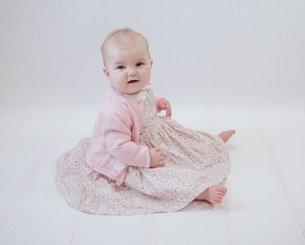 Small baby wearing pink sweater sitting on white floor with floral print dress. Early years pop up photo shoot