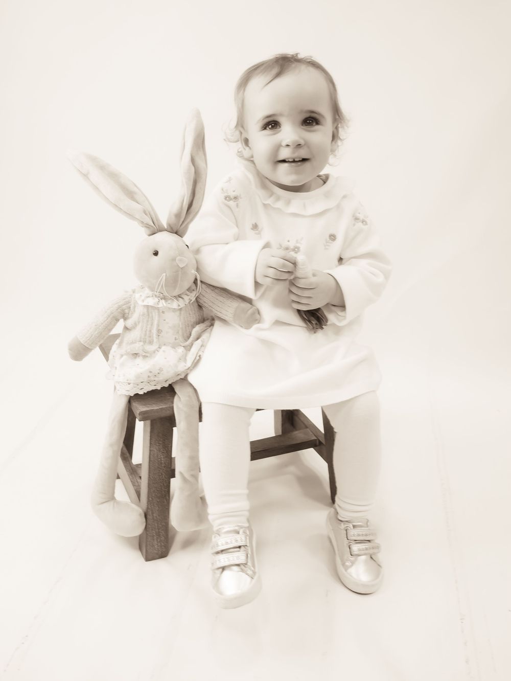 A toddler in a light dress and leggings sits on a wooden stool next to a stuffed bunny toy in a sepia-toned photo.