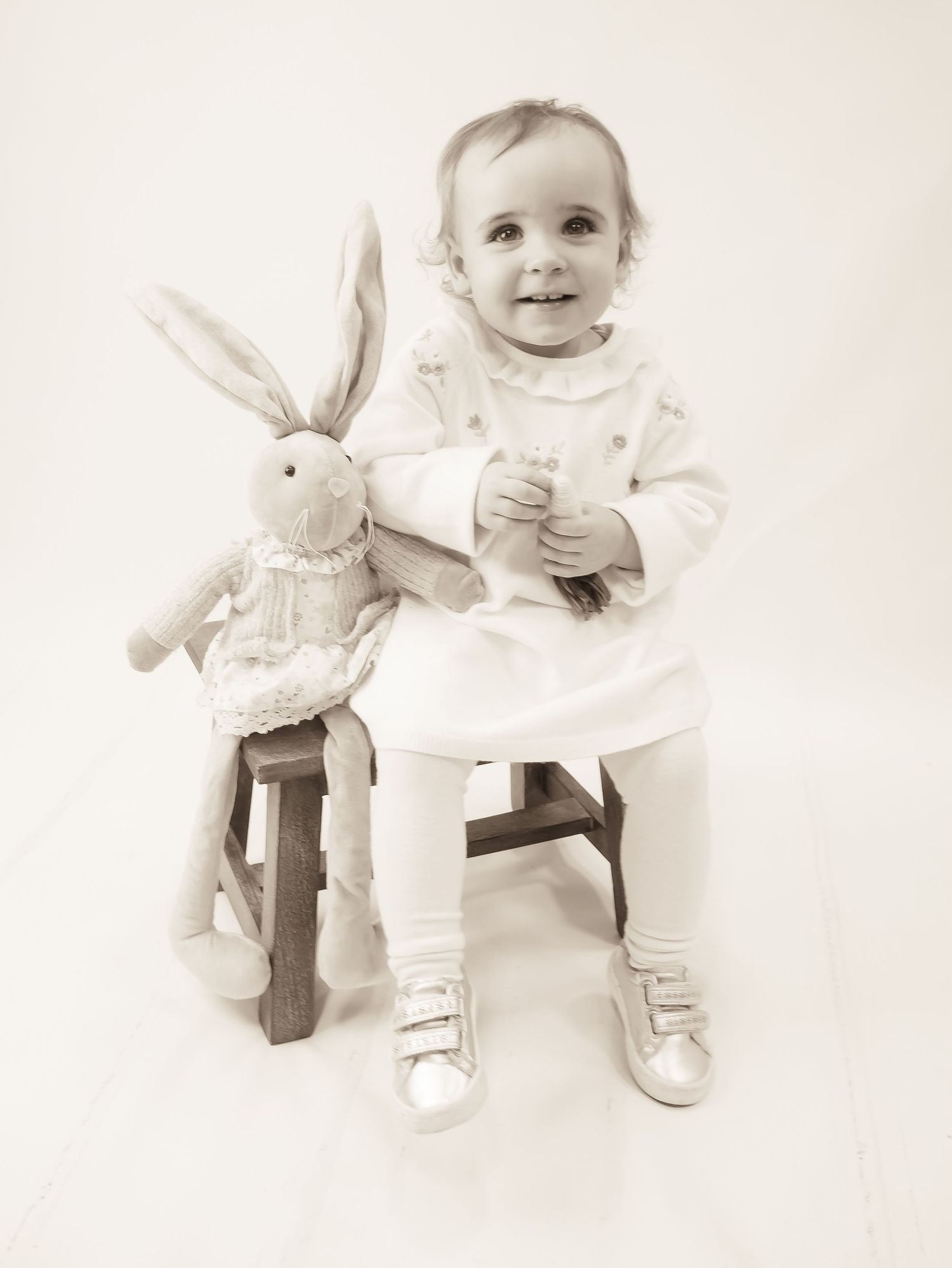 A toddler in a light dress and leggings sits on a wooden stool next to a stuffed bunny toy in a sepia-toned photo.
