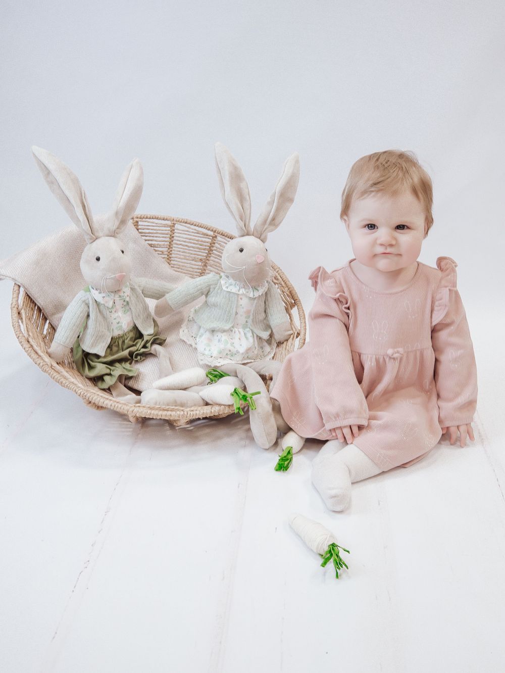 A baby in a pink outfit sits beside a basket containing two white stuffed bunny toys and floral accents. Easter photoshoot