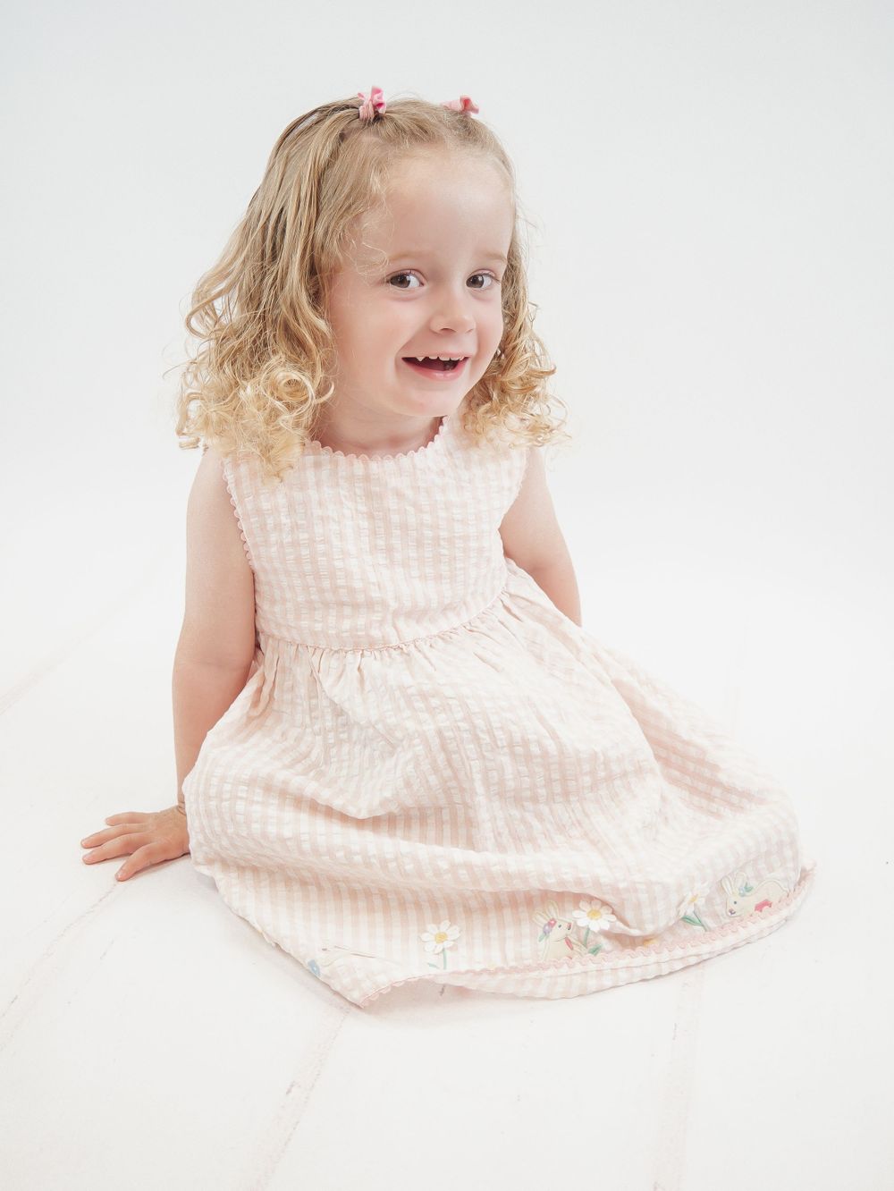 Young curly-haired child wearing a pale pink gingham dress sits smiling on a white background. School photos