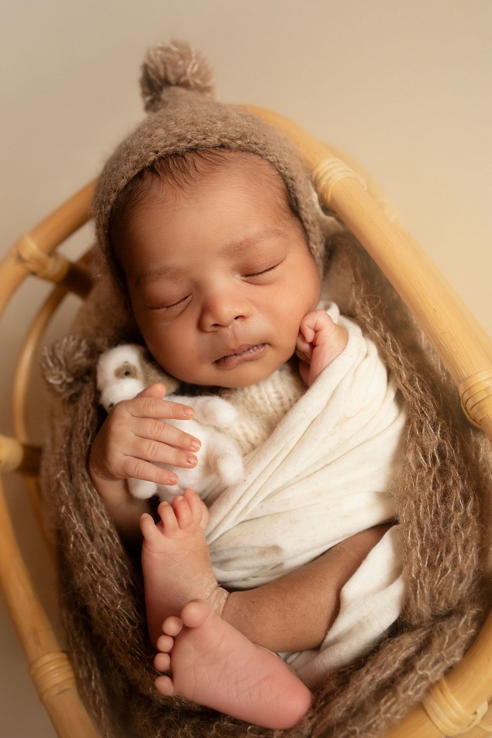 Sleeping newborn wrapped in white fabric posed in wooden bowl with brown textured blanket.