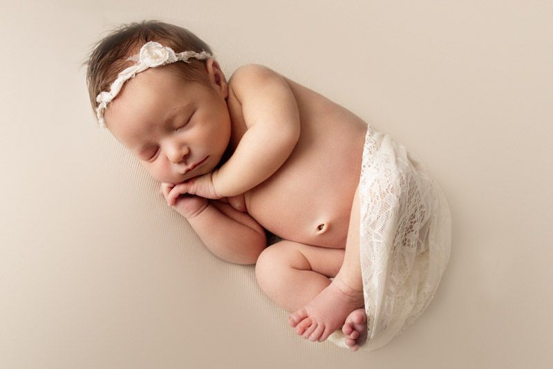 Sleeping newborn with lace headband draped in white fabric on beige backdrop.
