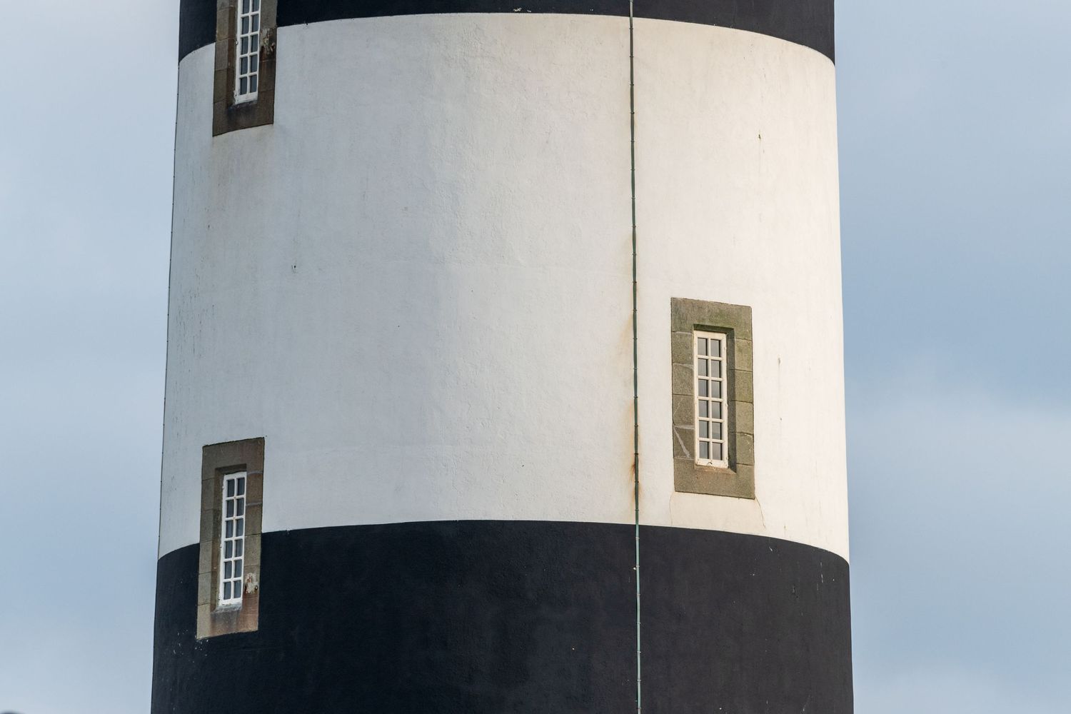 Close-up view of a black and white striped lighthouse tower with small rectangular windows against a cloudy sky.