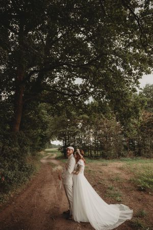 Wedding couple poses on rustic dirt road surrounded by lush green trees.