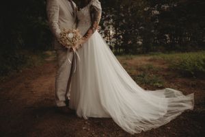Elegant wedding dress train flows on dirt path as couple embraces in moody lighting.