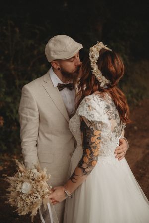 Wedding couple in beige suit and lace dress share tender moment with dried flower bouquet.