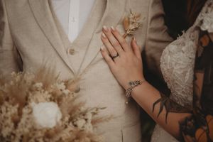 Close-up of couple's hands with wedding rings and dried floral bouquet against beige suit.