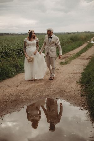 Wedding couple walking on dirt road with their reflection in puddle on overcast day.
