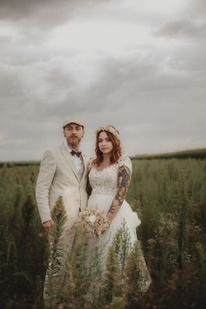 Newlyweds standing in field with cloudy sky and rustic bohemian wedding attire.