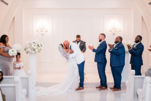 Wedding party poses in elegant white chapel with groomsmen in blue suits and bride in flowing white gown.