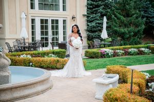 Elegant white lace wedding dress photographed in front of luxurious estate with manicured garden and fountain.
