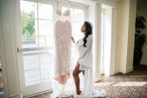 A blush pink wedding gown hangs by a bright window while someone in a white robe reaches towards it.