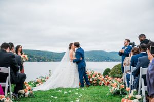 A scenic outdoor wedding ceremony takes place on a hilltop overlooking a serene lake with floral arrangements lining the aisle.