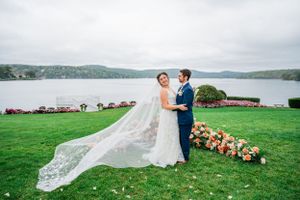 A wedding couple stands on a green lawn overlooking a serene lake with floral decorations and a long flowing veil.