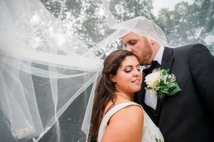 A romantic newlywed couple embrace on their wedding day with a flowing veil and white bouquet against a dreamy outdoor setting.