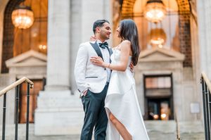 Couple in wedding attire embracing and smiling on elegant building steps, groom in white tuxedo, bride in white gown in New York City.
