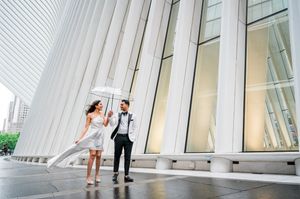Couple in wedding attire sharing umbrella outside modern white architectural building in New York City.