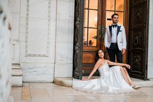 Bride in white gown sits on marble steps while groom in white tuxedo jacket stands beside ornate wooden doors in New York City