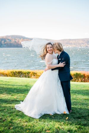 A romantic wedding couple embraces on a grassy lawn overlooking a scenic lake and mountains on their special day.