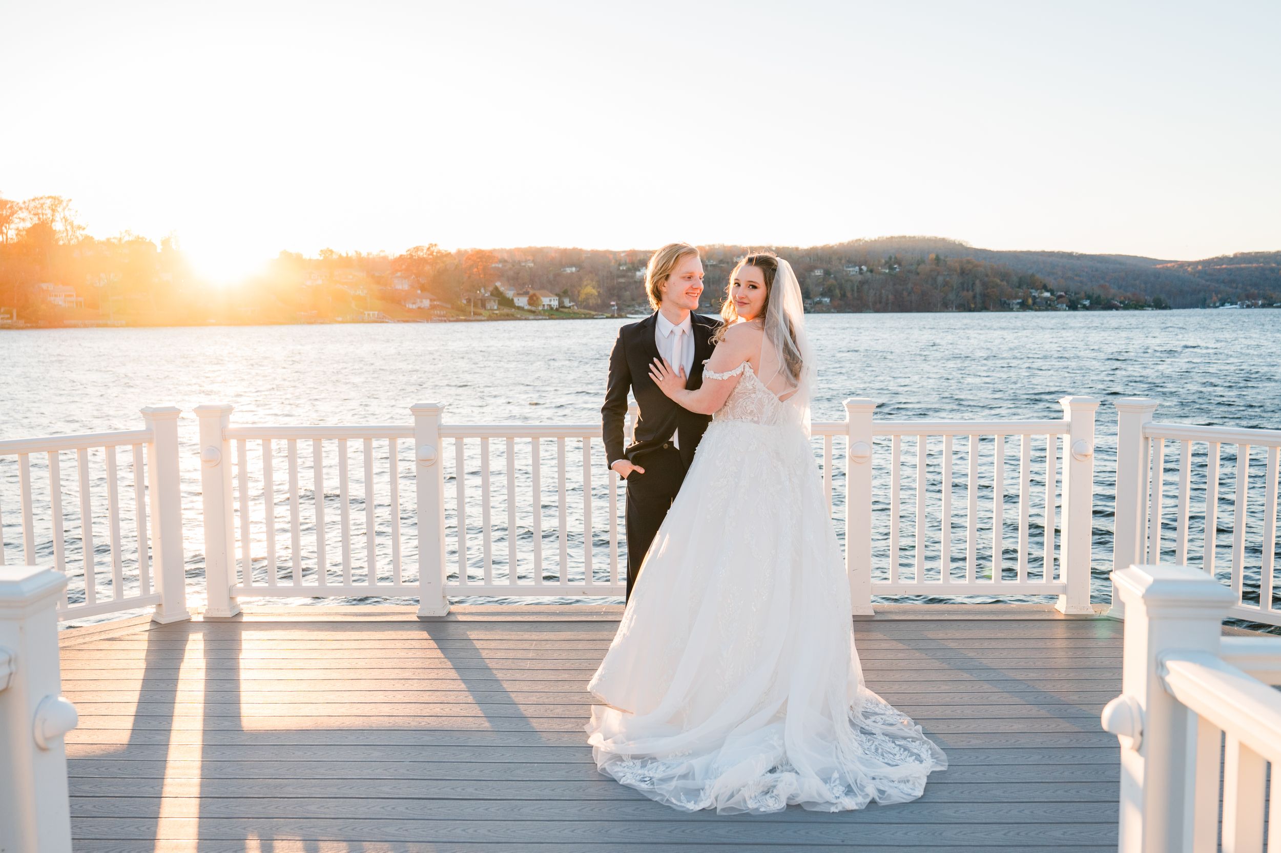 A couple shares an embrace on a waterfront dock during sunset as light glimmers across the lake.
