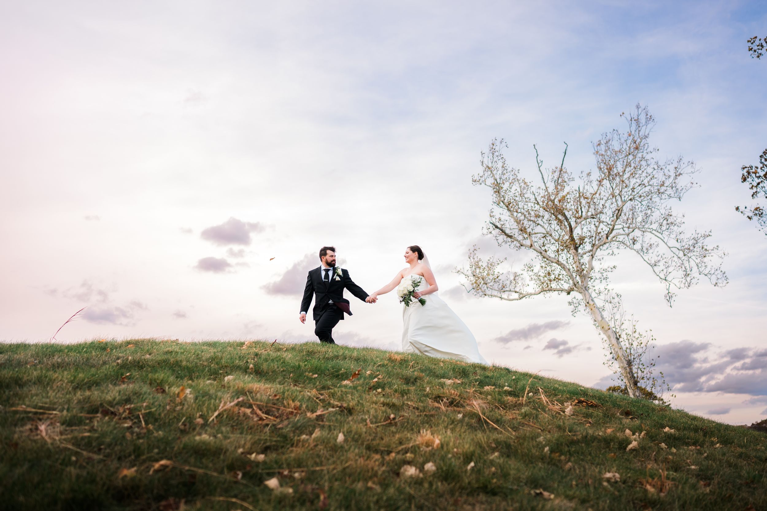 Bride and groom holding hands running on a grassy hill, wedding veil flowing in the wind against a cloudy sky at Great River Golf Club.