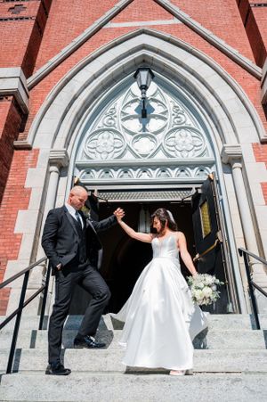 A couple in wedding attire celebrates on the steps of a historic red brick church with Gothic arched doorway.