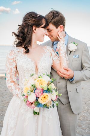 A romantic beach wedding scene with couple in lace gown and grey suit holding a vibrant pastel bouquet.