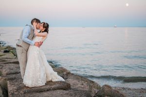 A romantic couple shares a kiss at sunset on rocky coastal shore with calm ocean waters and soft pink sky  at the Pavilions at Penfield