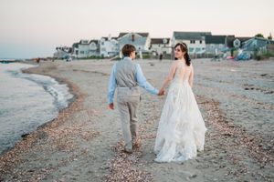 A couple walks hand in hand along a pebbly beach at dusk with coastal homes in the background.